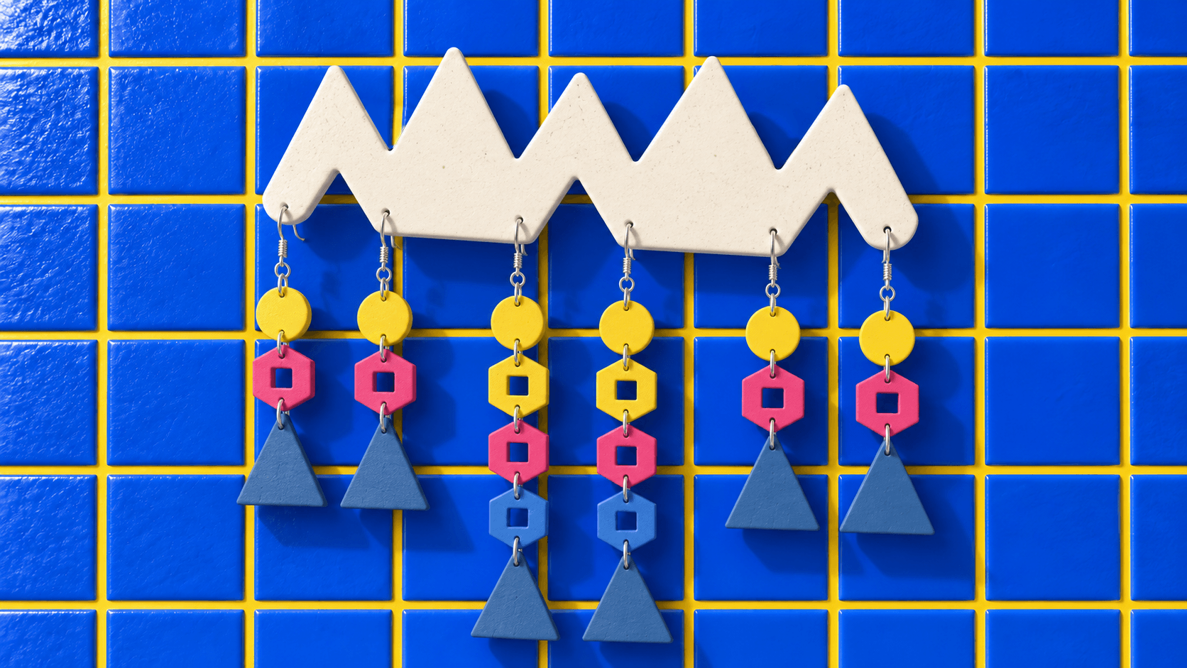 Cream Go-Go zigzag wall display holding three complete pairs of linked earrings with square windows, against blue tiles.